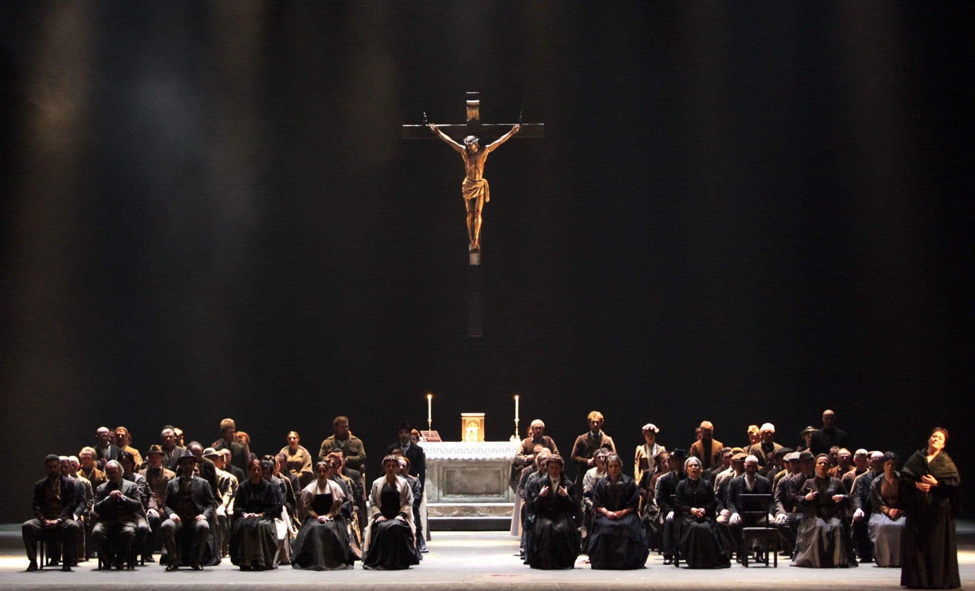 Scena terza. La scena si svolge in chiesa, con la funzione solenne che segue la processione pasquale. Il centro del palcoscenico è occupato da circa 60 sedie, disposte metà a sinistra e metà a destra, lasciando libero un corridoio centrale. Vi sono seduti i paesani, donne e uomini, vestiti con abiti semplici sulle tonalità del nero, del grigio e del marrone. Mamma Lucia è seduta in prima fila, sulla destra. Alla estrema destra, in piedi, Santuzza. Dietro le sedie, altri paesani sono in piedi. Perpendicolare al corridoio centrale, un altare di marmo con un leggìo, due ceri accesi, un calice liturgico di colore oro e un tabernacolo dello stesso colore. Sopra all’altare, sostenuto al soffitto da due corde, un grande crocifisso di legno, con il corpo di Gesù illuminato da una intensa luce calda, tendente al giallo. Il fondale del palcoscenico è nero.