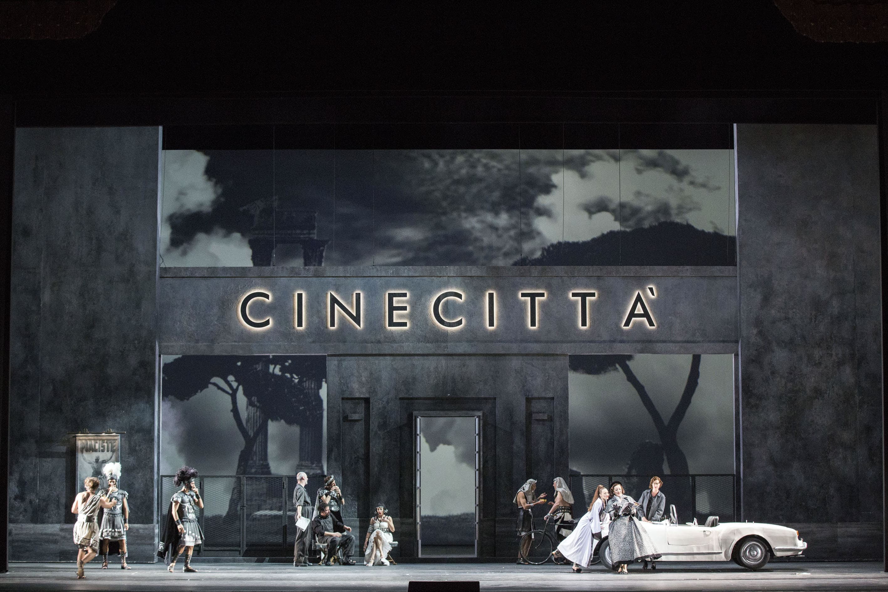 Primo atto. La scena si svolge presso l’ingresso di Cinecittà, rappresentato da una parete di colore grigio con, al centro, la scritta in stampatello “cinecittà”, illuminata. La parete ha delle aperture centrali che lasciano intravedere il fondale, su cui sono proiettati, in bianco e nero, dei pini e, a sinistra, due colonne che rappresentano i fori romani. Sulla sinistra della parete è affisso un poster con la scritta “Maciste”. Di fronte alla parete, i figuranti usciti da un set cinematografico vestiti da centurioni, romani e romane conversano tra loro. All’estrema destra del palcoscenico, Norina, vicino alla sua macchina modello Spider, di colore bianco, mostra ad una donna la pagina di una rivista che tiene aperta. È vestita con un cappotto maculato, bianco e nero, lungo fino ai piedi, stretto in vita da una cintura nera. Indossa dei guanti neri e un cappellino dello stesso colore.