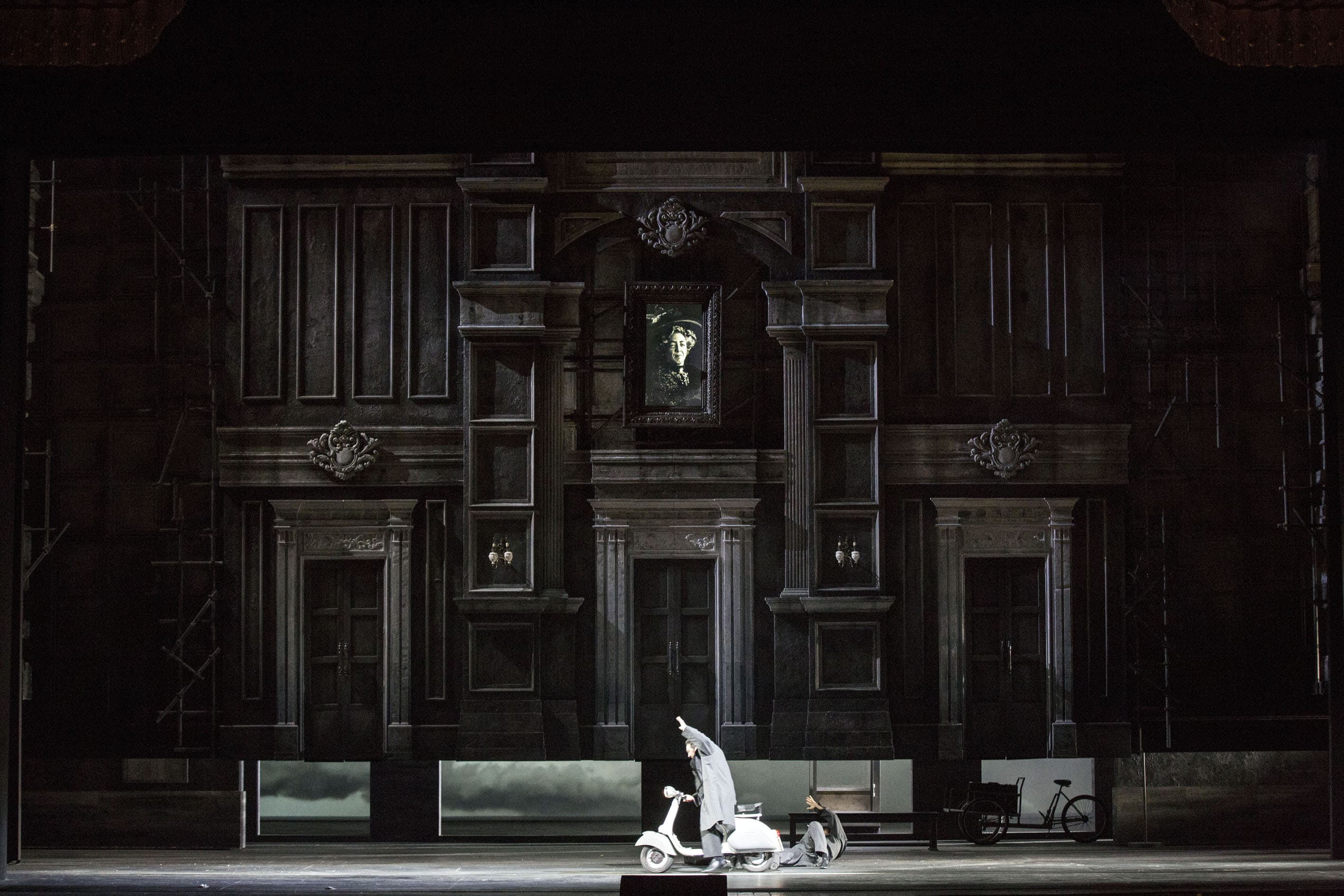 Secondo atto. La scena si svolge nella casa di Don Pasquale, con la parete di legno scuro, qui leggermente sollevata e lasciando intravedere l’atrio della stazione ferroviaria. Al centro del palcoscenico, una vespa che richiama quella del film Vacanze Romane. Un uomo è alla guida, con il braccio sinistro alzato, e un altro, il passeggero, è appena caduto a terra. I due sono vestiti con lunghi impermeabili di colore grigio. 