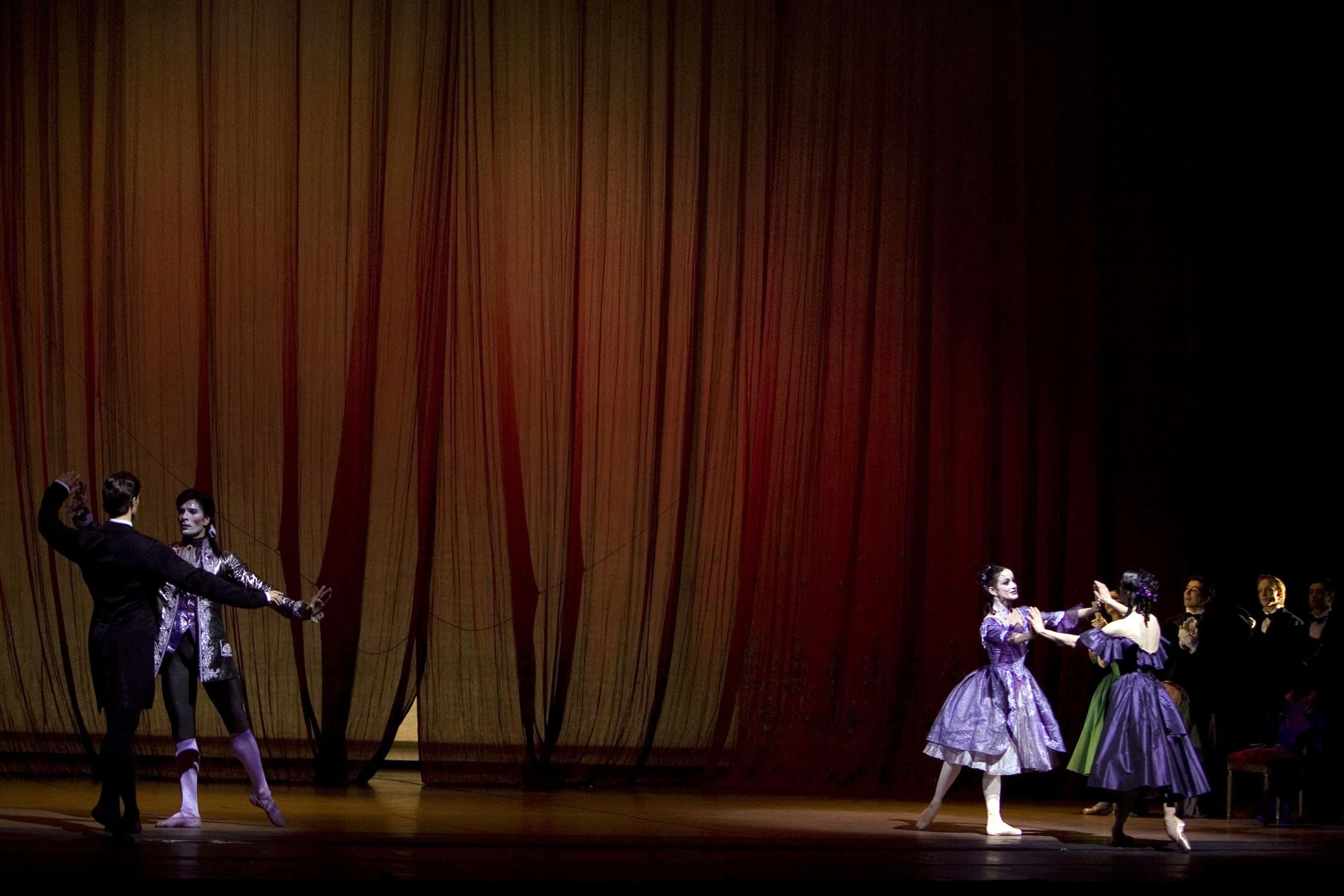Primo atto. La scena si svolge presso il Théâtre-des-Variétés di Parigi, dove si è appena concluso il balletto Manon Lescaut. Sul fondo del palcoscenico, un grande sipario dipinto, di tulle, di colore rosso. Al centro, sulla sinistra, Armand è di spalle al pubblico, di fronte a Des Grieux, il protagonista maschile del balletto, mentre sulla destra Marguerite è davanti a Manon e le loro mani si sfiorano. Armand indossa un frac nero mentre Marguerite un abito viola, lungo fino a metà polpaccio. Il corpetto ha un ampio scollo che lascia scoperte le spalle, mentre la gonna è a balze. All’estrema destra del palco, alcune poltroncine di velluto rosso su cui siedono gli spettatori, tra cui Prudence, amica di Marguerite. Gli uomini indossano dei frac di colore nero, dei guanti bianchi e applaudono verso il centro del palco. Prudence indossa un abito lungo, di colore verde, e stringe in mano un ventaglio.