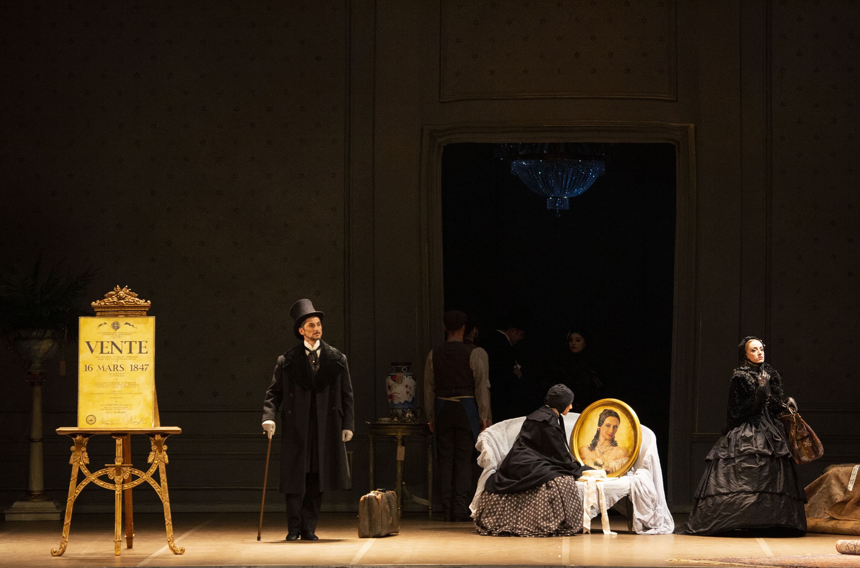 Prologo. La scena si svolge in una delle stanze dell’appartamento ormai dismesso di Marguerite Gautier. Il fondale è in tulle, dipinto a simulare una parete con boiserie. Al centro del fondale è presente il varco di una porta (aperta, senza battenti) che rappresenta l’ingresso della stanza. Dietro si intravede un lampadario coperto con dei teli. A sinistra dell’ingresso, un tavolino con sopra un vaso da fiori. A sinistra della stanza, un cavalletto di colore oro mostra il manifesto “Vente 16 mars 1847” (in francese, vendita 16 marzo 1847). Al centro, Monsieur Duval è in piedi, vicino ad una valigia, appoggiata a terra. Indossa un elegante cappotto nero, doppiopetto, con il collo di pelliccia nera, un cappello a cilindro, sempre nero, e dei guanti bianchi. Si appoggia ad un bastone da passeggio. A destra di Duval, un divanetto coperto da un telo bianco, su cui è appoggiato un ritratto di Marguerite in una grande cornice ovale intarsiata. Sul divanetto vi è seduta Nanine, cameriera di Marguerite, che, dando le spalle al pubblico, osserva il ritratto. Indossa un abito lungo con la gonna a campana, di colore marrone e decori bianchi. Sopra alle spalle indossa una lunga mantella nera e in testa una cuffietta dello stesso colore, legata sotto al mento. A destra di Nanine, una dama, in piedi, stringe in mano un elegante borsone. Indossa un abito nero, lungo fino ai piedi, e dei guanti di pizzo, sempre neri; in testa, una cuffietta dello stesso colore, legata sotto al mento. 